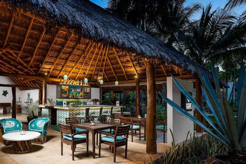 Tropical beachfront bar with thatched roof, wooden furnishings, and turquoise accent seating