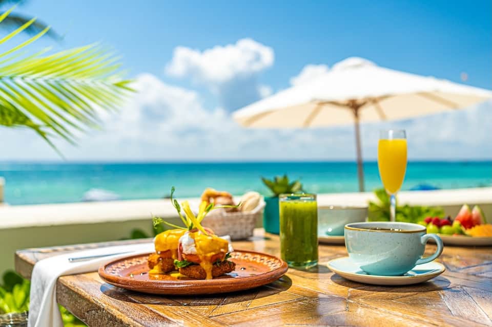 Beachfront breakfast setting with tropical fruit plate, coffee, and ocean view under umbrella
