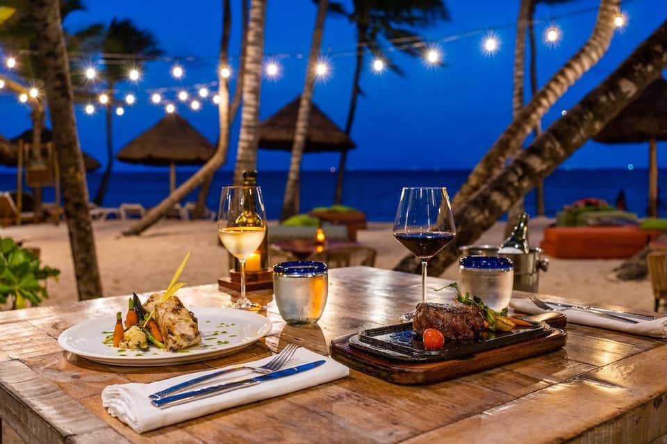 Beachfront fine dining table at night with wine glasses, plated entrees, and ocean view framed by palm trees with string lights