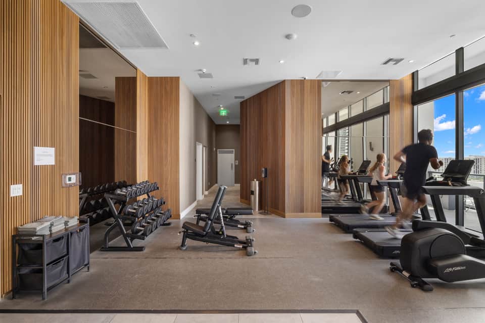 Modern hotel fitness center with cardio equipment, dumbbells, wooden wall panels, and floor-to-ceiling windows