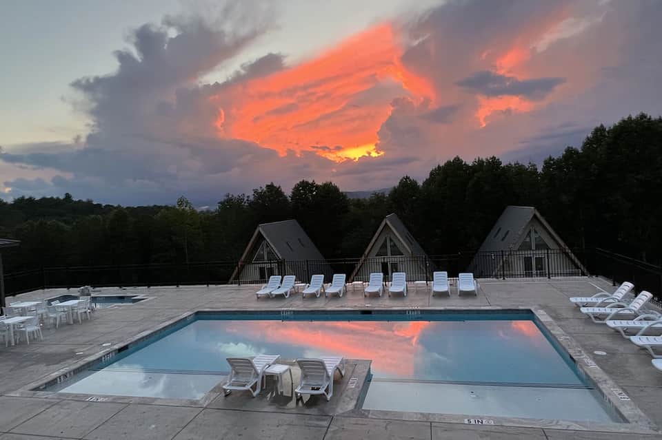 Resort pool at sunset with white lounge chairs, A-frame cabins, and dramatic orange sky