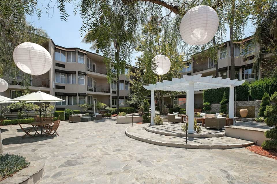 Resort courtyard with white pergola, lounge seating, fire pit, weeping willow trees, and modern apartments