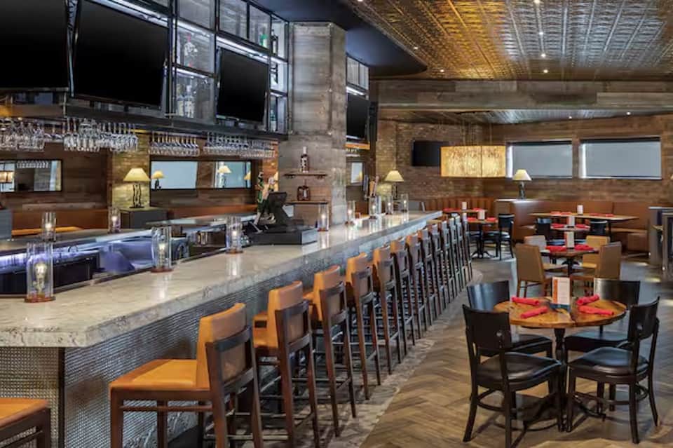 Upscale restaurant interior with marble bar counter, orange seating, wood accents, and multiple television screens.