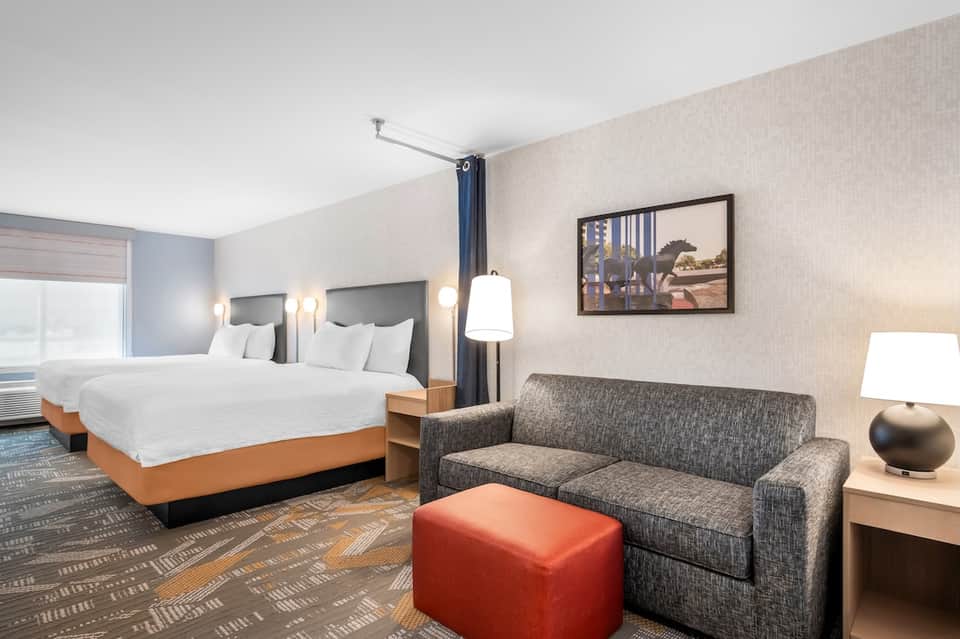 Hotel guest room with two queen beds, gray sofa, red ottoman, and contemporary décor
