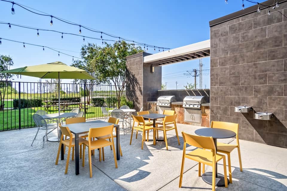 Outdoor patio with yellow chairs, tables, gas grills, green umbrella, and lawn