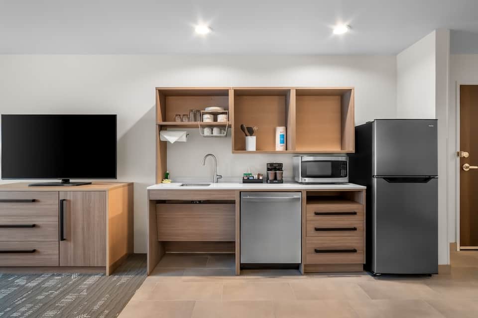 Hotel room kitchenette with wood cabinetry, stainless appliances, open shelving, and flat-screen TV