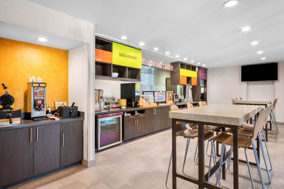 Contemporary hotel breakroom with yellow accent wall, dark cabinetry, high-top table, and colorful shelving