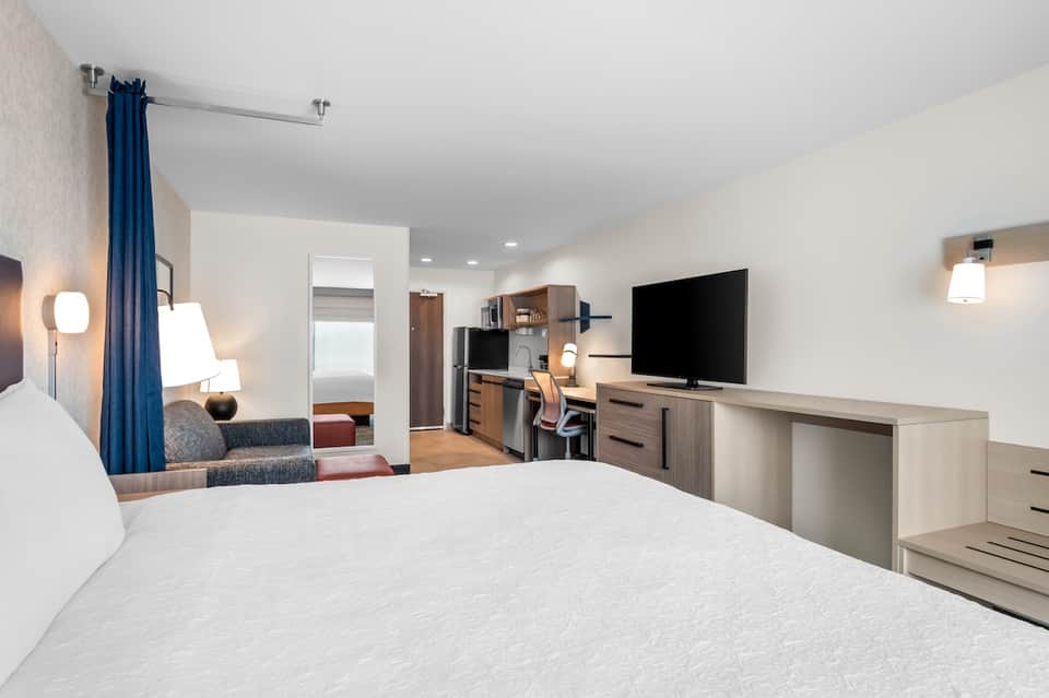 Hotel guest room with king bed, work desk, flat-screen TV, blue curtains, kitchenette, and warm contemporary decor