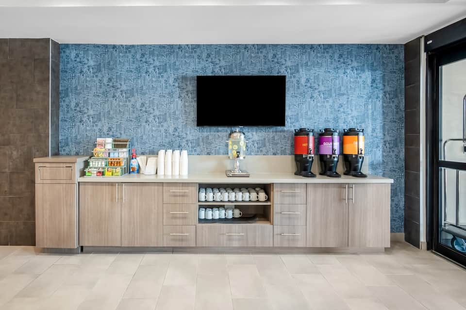 Hotel lobby beverage station with blue tile wall, TV, coffee makers, and supply storage