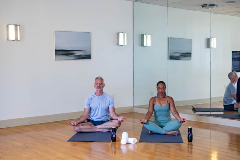 Yoga studio with two people meditating on mats, mirror wall, wooden floor, and minimalist coastal décor