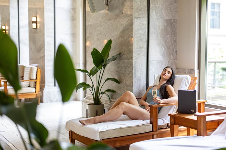 Woman relaxing on lounge chair in spa area with marble walls, potted plants, and champagne glass