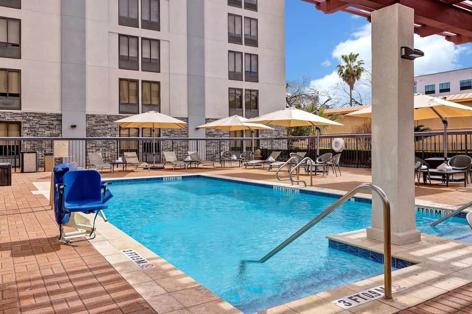 Hotel courtyard pool with blue water, tan umbrellas, lounge chairs, and multi-story building with stone accents.