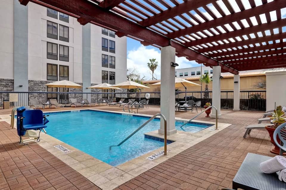 Resort pool with covered pergola, blue water, loungers, multi-story building, and palm trees