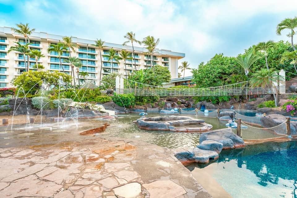 Resort pool area with natural rock features, blue water, tropical plants, and multi-story hotel building
