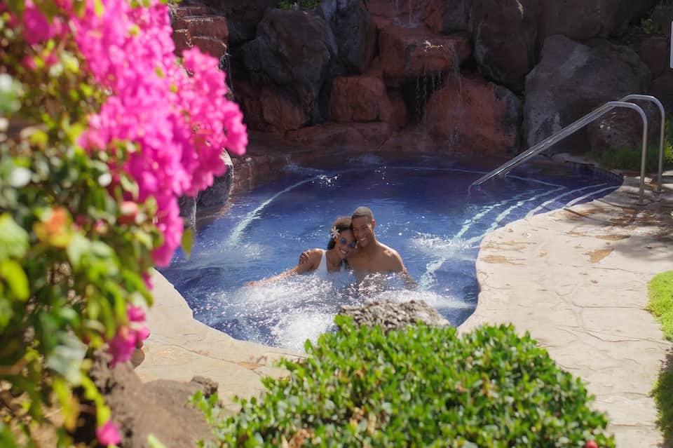 Two guests enjoying a hot spring pool surrounded by pink flowering plants and rocky landscape