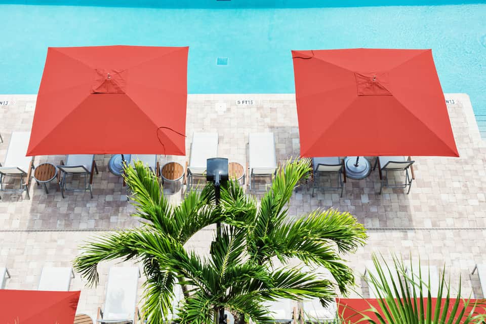 Aerial view of bright turquoise pool with red umbrellas, white lounge chairs, and palm trees