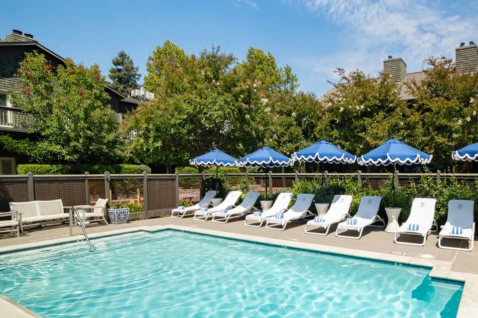 Resort pool with blue umbrellas, lounge chairs, wooden fence, and residential buildings