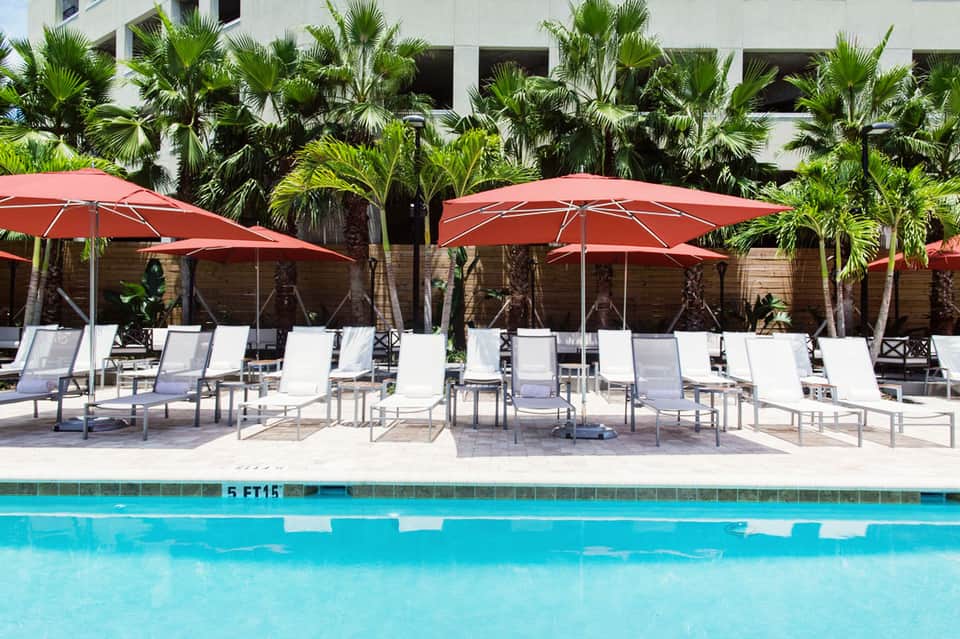 Resort pool area with white lounge chairs, red umbrellas, and palm trees lining the deck