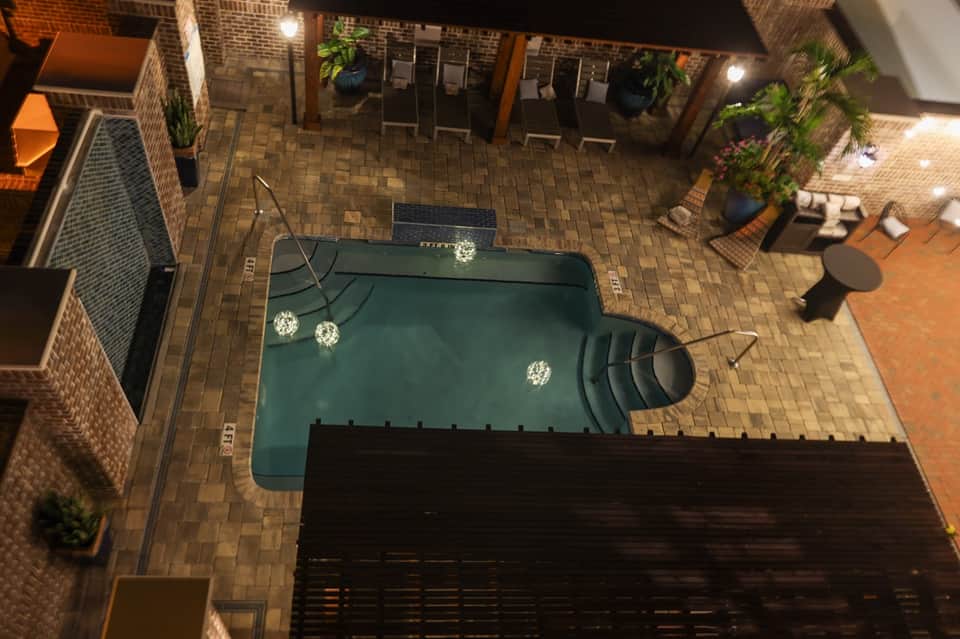 Aerial view of small hotel pool at night with brick courtyard and lounge chairs