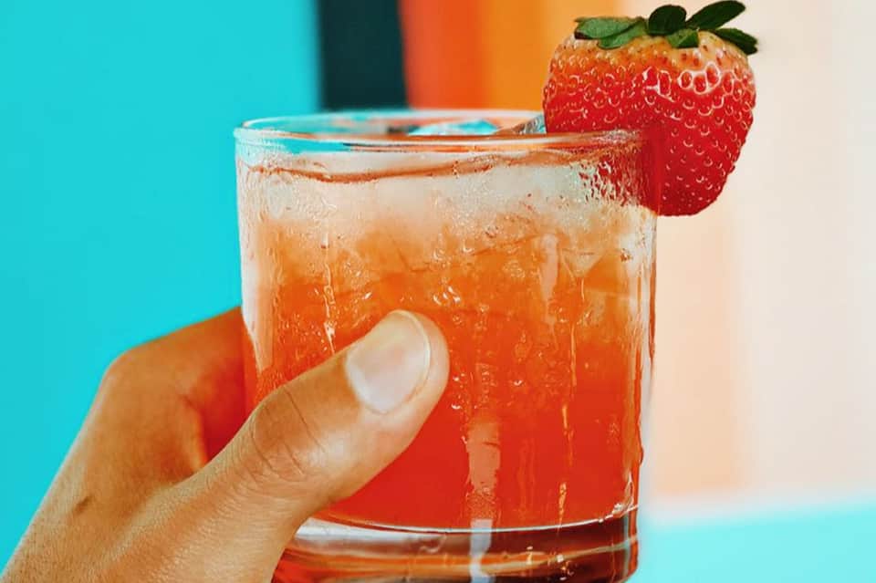 Strawberry-garnished cocktail with orange liquid and ice in vibrant turquoise setting