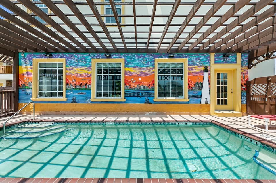 Indoor pool with geometric shadow patterns and vibrant yellow building with painted ocean and sunset mural backdrop