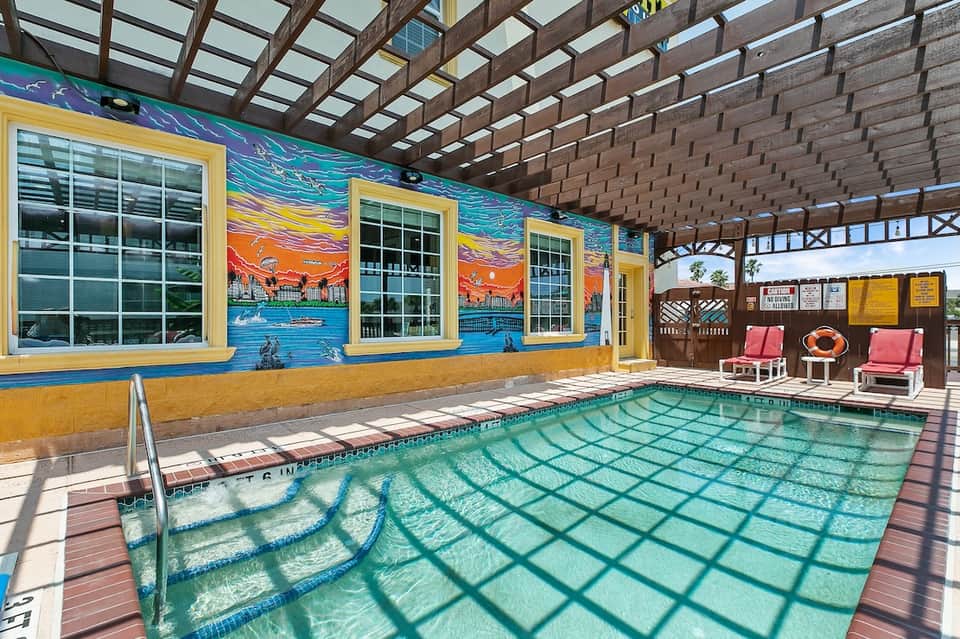 Indoor heated pool with colorful mural, yellow trim, wooden pergola ceiling, and lounge chairs poolside