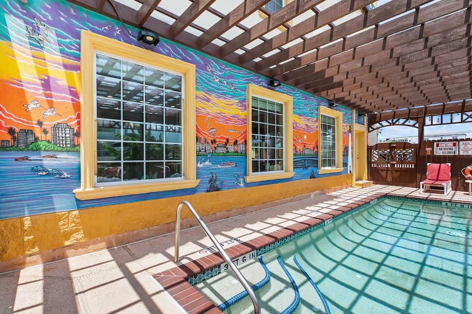 Indoor heated pool with colorful mural artwork, pergola ceiling, and lounge chairs