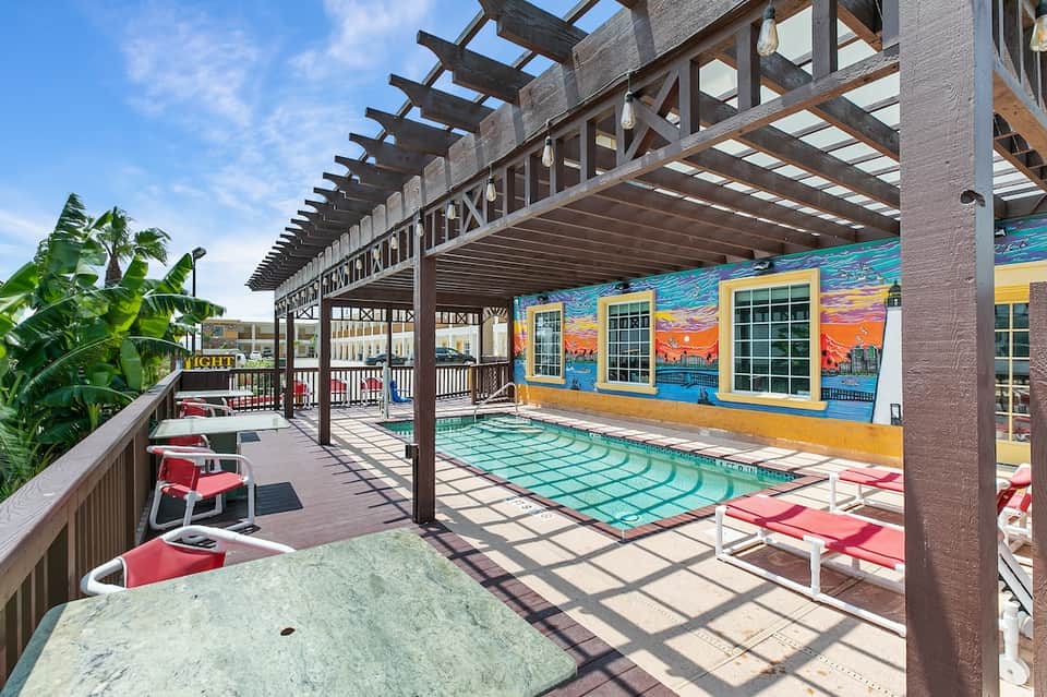 Covered pool deck with dark wooden pergola, turquoise water, colorful mural, and palm vegetation