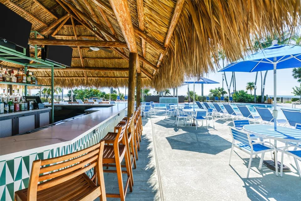 Tiki-style beach bar with thatched roof, wooden chairs, blue umbrellas, and waterfront palm tree setting