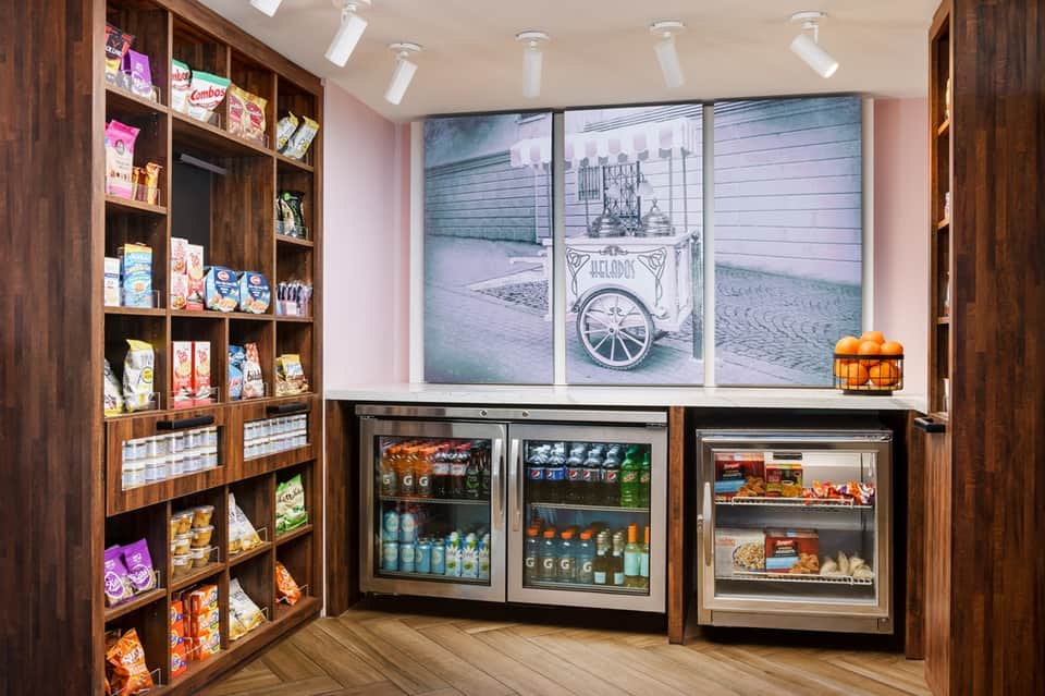 Modern convenience shop with wood shelving displaying snacks and beverages, refrigerated cases, and vintage cart mural