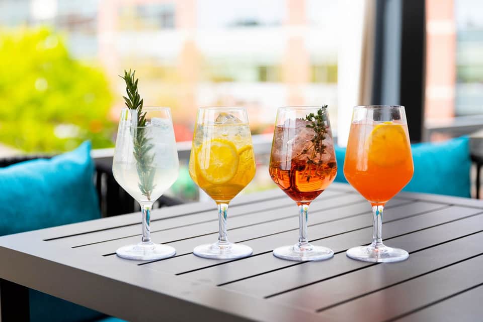 Four colorful cocktails with garnishes lined up on outdoor table with city buildings in background