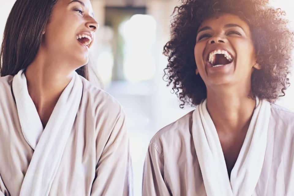 Two women laughing together wearing spa robes in bright, airy wellness setting