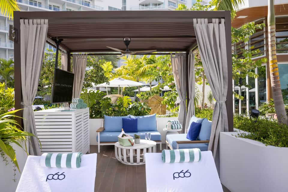 Outdoor lounge pavilion with gray curtains, blue cushioned seating, and tropical vegetation