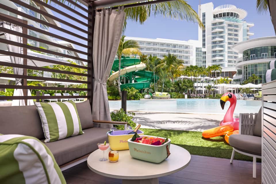 Tropical pool cabana with striped shade structure, water slide, and modern resort buildings