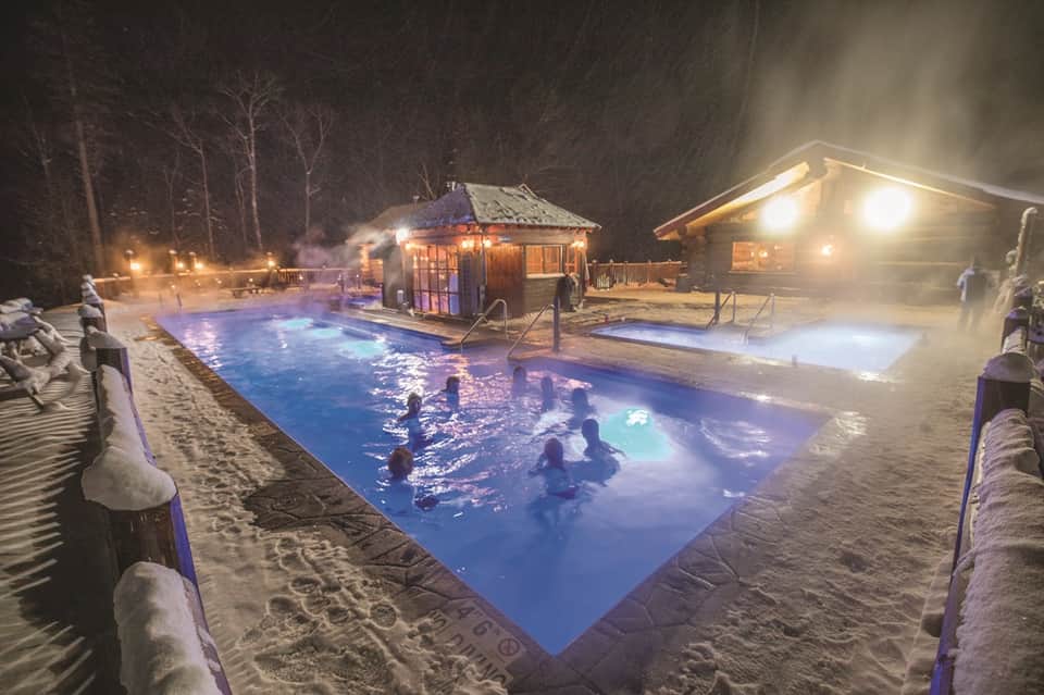 Outdoor hot spring pool at night with illuminated water, wooden cabins, and snow surrounding area