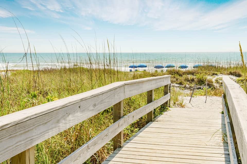 Wooden boardwalk leading to sandy beach with dune grass and blue umbrellas beyond the shoreline