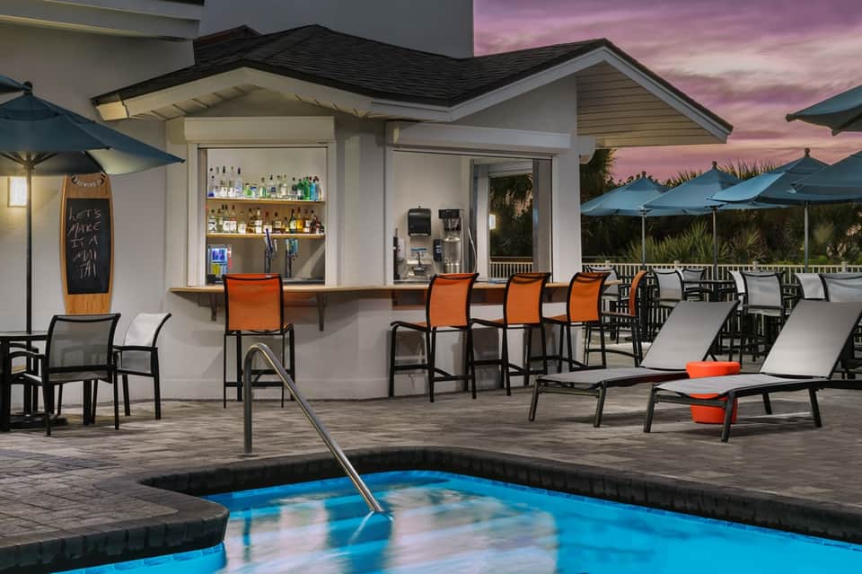 Pool deck at dusk with blue hot tub, bar station, orange lounge chairs, and teal umbrellas
