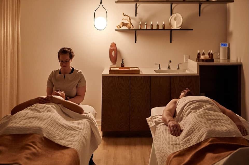 Spa treatment room with two massage tables and therapist preparing wellness service