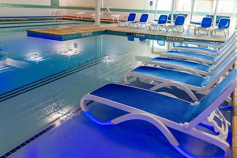 Indoor lap pool with lane markers, blue lounge chairs, diving board, and natural light from windows