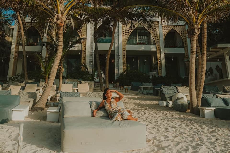 Beach club sand lounge area with white cushioned seating under arched architecture with palm trees