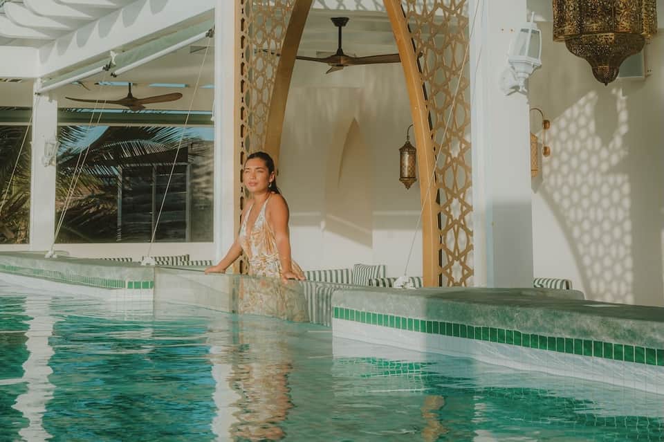 Woman in white dress standing in turquoise indoor pool with Moroccan-style doors and hanging lanterns