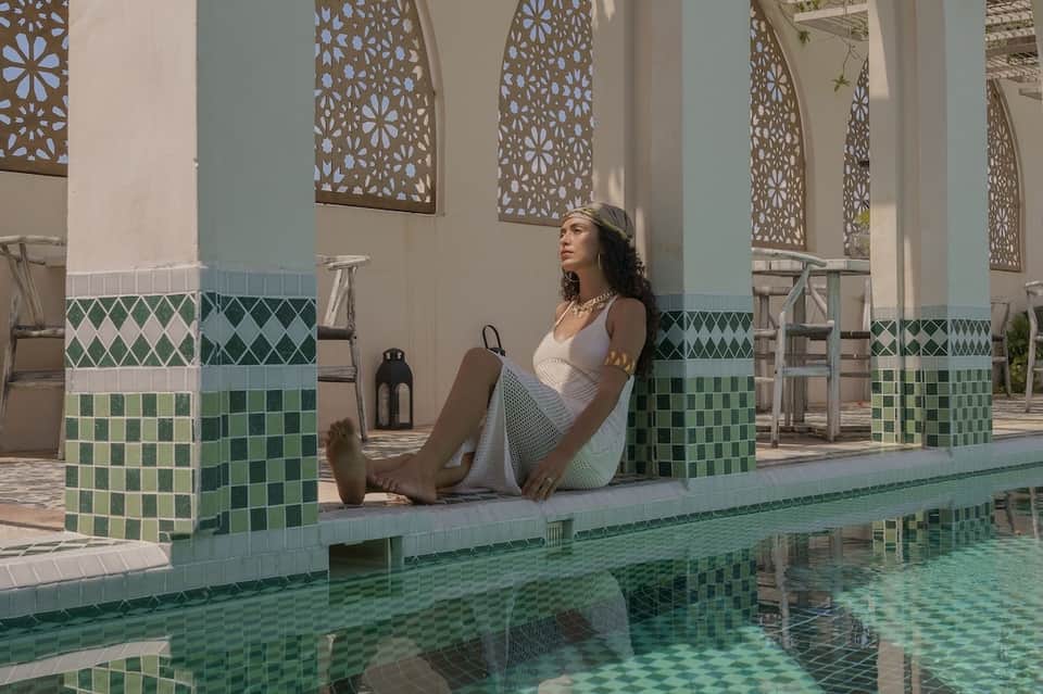 Woman in white dress lounging by decorative tiled pool with Moroccan-style architectural details