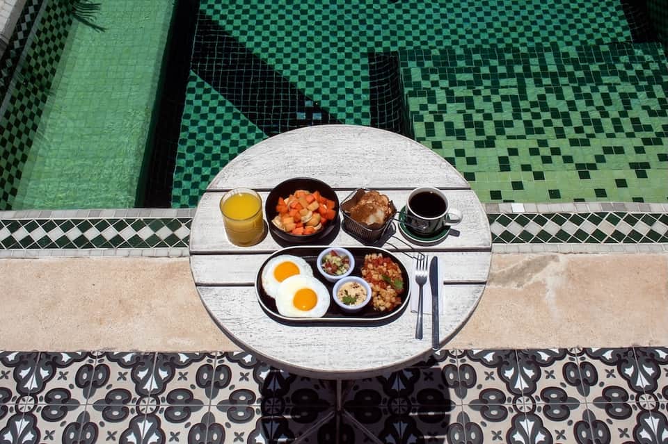 Breakfast tray by green tiled pool featuring eggs, fruit, pastries, juice, and coffee at waterside