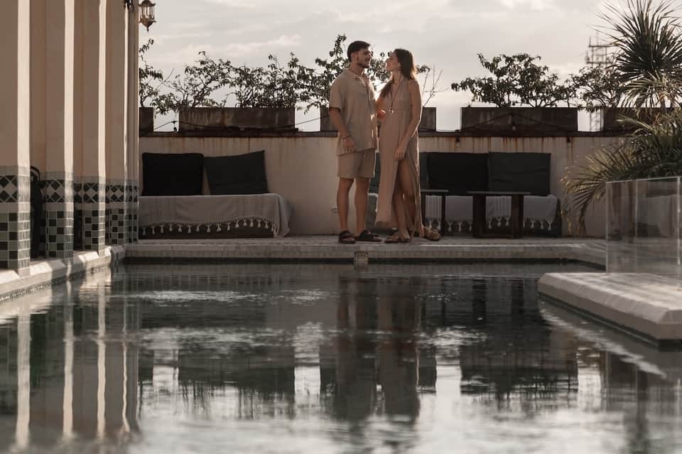 Couple standing on rooftop terrace overlooking infinity pool at dusk with potted plants and city views