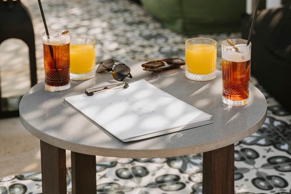 Outdoor poolside table with beverages, sunglasses, notepad, and patterned floor in natural sunlight