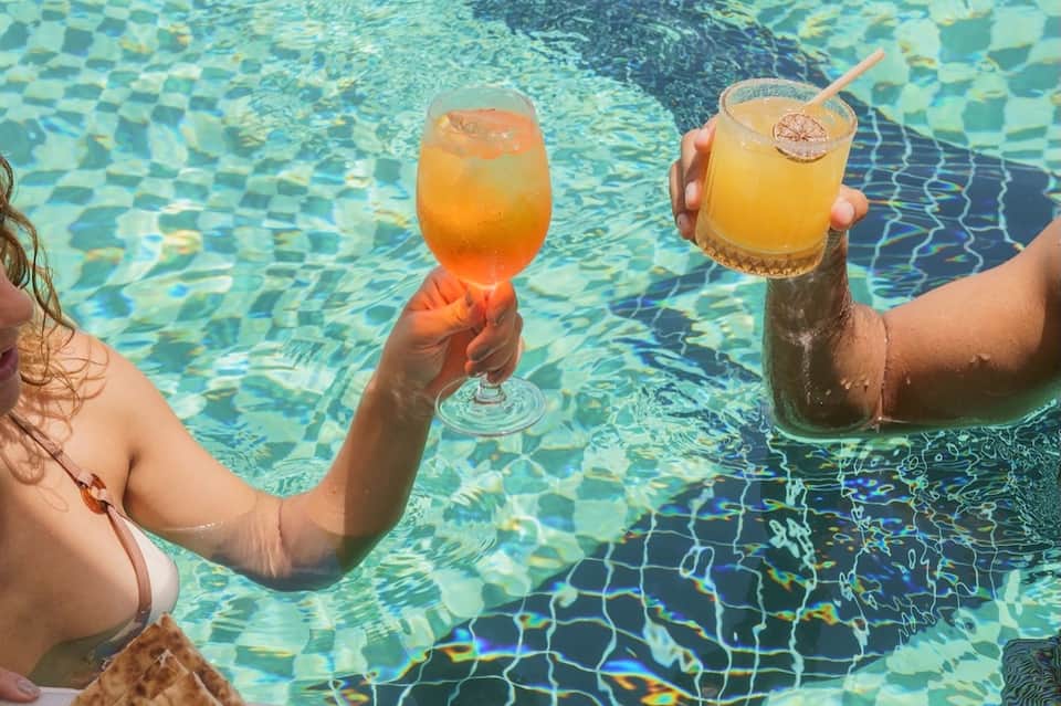 Guests holding colorful cocktails while swimming in clear turquoise pool with rippling water reflections