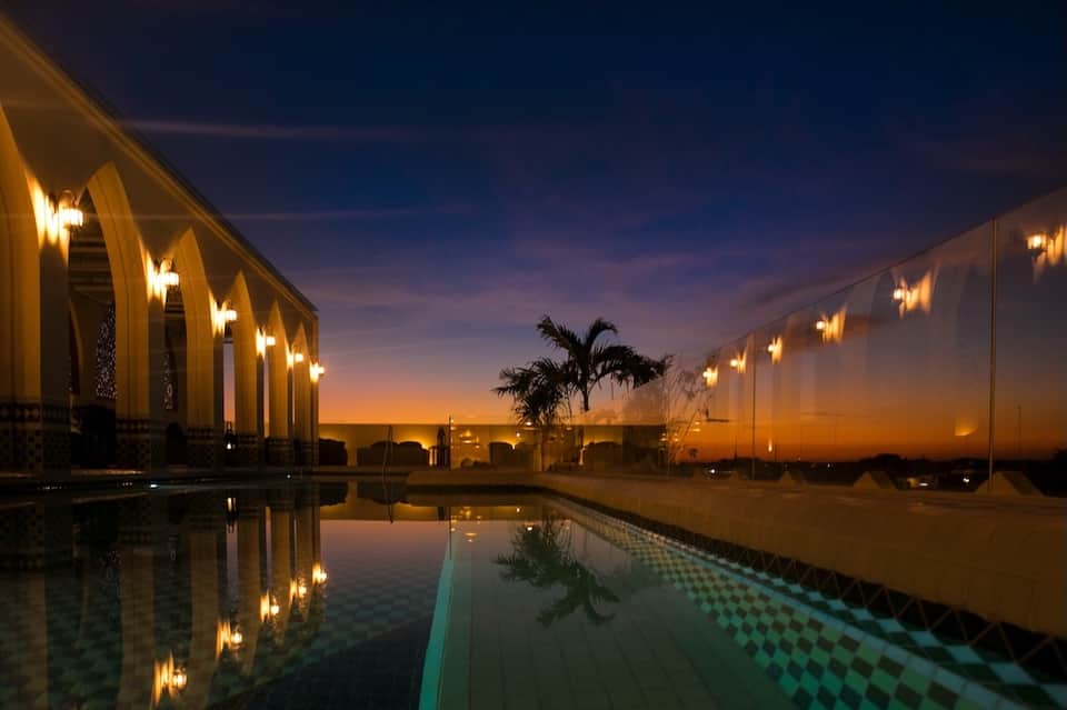 Illuminated resort pool and pavilion at dusk with palm trees reflected in still water at sunset