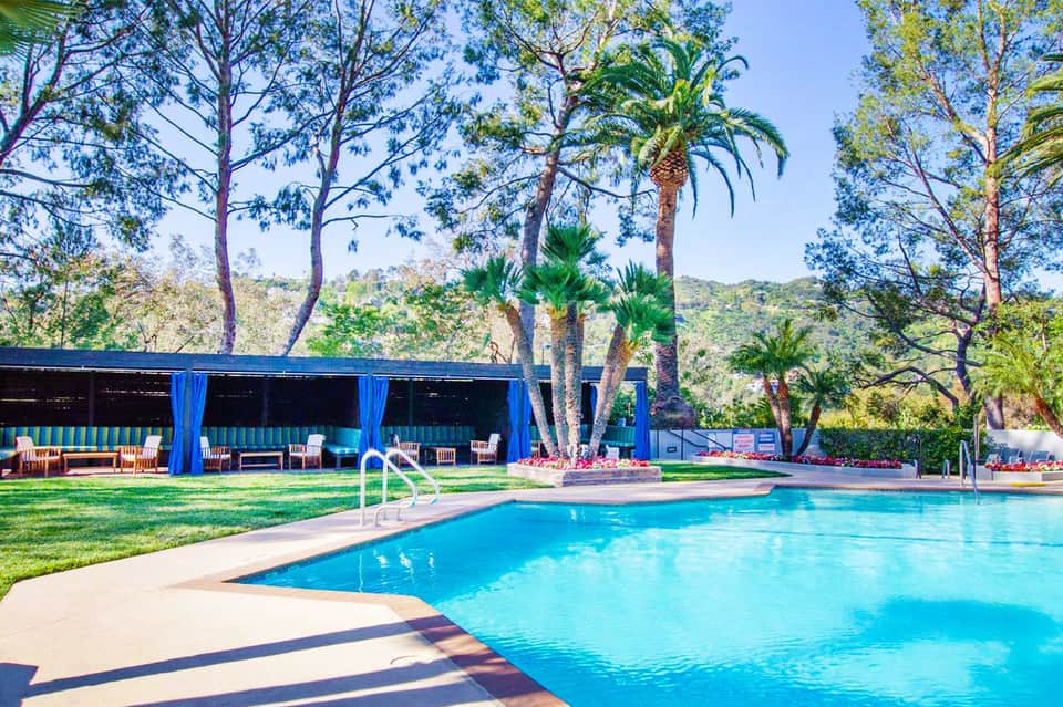 Resort pool surrounded by palm trees and tall pines with blue cabana structure and lounge chairs