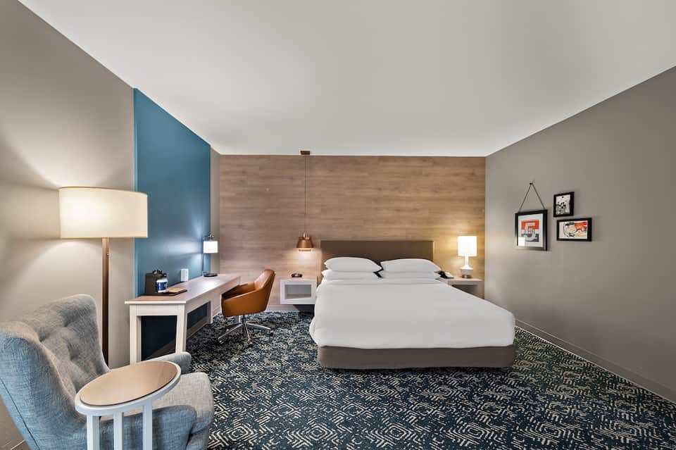 Modern hotel bedroom with blue accent wall, wood headboard, desk, lounge chair, and neutral decor