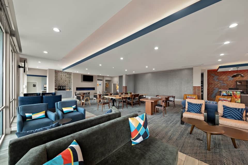 Spacious hotel lobby lounge with blue seating, exposed brick, fireplace, dining area, and colorful accent pillows